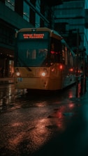 A yellow tram is moving along wet streets reflecting red and yellow lights, surrounded by modern urban buildings. The destination sign on the tram glows with the text 'Etihad Campus via Piccadilly'.