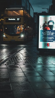 A city street at night with a wet reflective pavement. A bus is approaching with its headlights on, displaying the route number 47 to Manchester. To the right, there is a lit advertisement board promoting a donation campaign for the Royal British Legion Poppy Appeal.