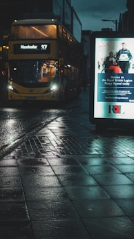 A city street at night with a wet reflective pavement. A bus is approaching with its headlights on, displaying the route number 47 to Manchester. To the right, there is a lit advertisement board promoting a donation campaign for the Royal British Legion Poppy Appeal.