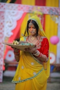 A woman dressed in a vibrant yellow saree with red and green accents stands holding a woven basket filled with bananas. She has an intricate nose ring and necklace, and her forehead is marked with orange sindoor. Behind her are colorful drapes in red and yellow, adorned with floral patterns.