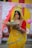 A woman dressed in a vibrant yellow saree with red and green accents stands holding a woven basket filled with bananas. She has an intricate nose ring and necklace, and her forehead is marked with orange sindoor. Behind her are colorful drapes in red and yellow, adorned with floral patterns.