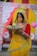 A woman dressed in a vibrant yellow saree with red and green accents stands holding a woven basket filled with bananas. She has an intricate nose ring and necklace, and her forehead is marked with orange sindoor. Behind her are colorful drapes in red and yellow, adorned with floral patterns.