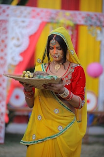 A woman dressed in a vibrant yellow saree with red and green accents stands holding a woven basket filled with bananas. She has an intricate nose ring and necklace, and her forehead is marked with orange sindoor. Behind her are colorful drapes in red and yellow, adorned with floral patterns.