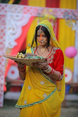 A woman dressed in a vibrant yellow saree with red and green accents stands holding a woven basket filled with bananas. She has an intricate nose ring and necklace, and her forehead is marked with orange sindoor. Behind her are colorful drapes in red and yellow, adorned with floral patterns.