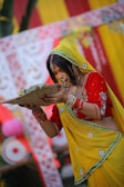 A woman in traditional Indian attire is participating in a cultural ceremony. She is wearing a vibrant yellow and red saree with intricate embroidery and accessories. Her hair is adorned with jewelry, and she holds a woven basket-like object with both hands. The background features colorful decorations, including red and yellow fabrics with floral designs.