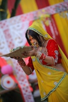 A woman in traditional Indian attire is participating in a cultural ceremony. She is wearing a vibrant yellow and red saree with intricate embroidery and accessories. Her hair is adorned with jewelry, and she holds a woven basket-like object with both hands. The background features colorful decorations, including red and yellow fabrics with floral designs.