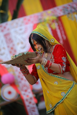 A woman in traditional Indian attire is participating in a cultural ceremony. She is wearing a vibrant yellow and red saree with intricate embroidery and accessories. Her hair is adorned with jewelry, and she holds a woven basket-like object with both hands. The background features colorful decorations, including red and yellow fabrics with floral designs.