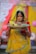 A woman dressed in traditional attire holds a woven basket containing fruits and other items. She wears a vibrant yellow saree with green and red accents, complemented by intricate jewelry, including bangles and a headpiece. The background features colorful fabrics and decorations, suggesting a festive or ceremonial setting.