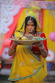 A woman dressed in traditional attire holds a woven basket containing fruits and other items. She wears a vibrant yellow saree with green and red accents, complemented by intricate jewelry, including bangles and a headpiece. The background features colorful fabrics and decorations, suggesting a festive or ceremonial setting.
