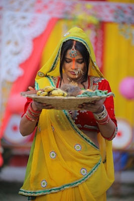 A woman dressed in traditional attire holds a woven basket containing fruits and other items. She wears a vibrant yellow saree with green and red accents, complemented by intricate jewelry, including bangles and a headpiece. The background features colorful fabrics and decorations, suggesting a festive or ceremonial setting.