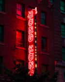 A neon sign with the words 'Hong Kong' in bright red letters is mounted vertically on the side of a brick building. The glowing sign is outlined by small white bulbs, which add extra illumination. Several windows are visible in the building, with some lights reflecting in them. The scene is set in the evening, creating a vibrant and slightly nostalgic urban atmosphere.