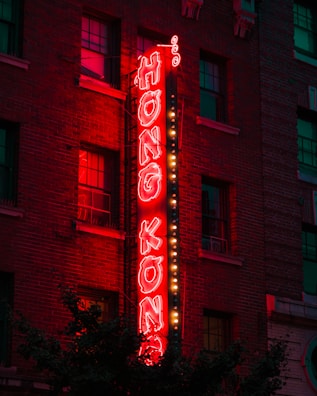 A neon sign with the words 'Hong Kong' in bright red letters is mounted vertically on the side of a brick building. The glowing sign is outlined by small white bulbs, which add extra illumination. Several windows are visible in the building, with some lights reflecting in them. The scene is set in the evening, creating a vibrant and slightly nostalgic urban atmosphere.