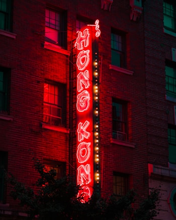 A neon sign with the words 'Hong Kong' in bright red letters is mounted vertically on the side of a brick building. The glowing sign is outlined by small white bulbs, which add extra illumination. Several windows are visible in the building, with some lights reflecting in them. The scene is set in the evening, creating a vibrant and slightly nostalgic urban atmosphere.