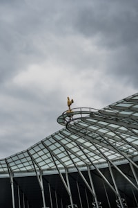 A modern architectural structure with a steel and glass roof design, topped with a golden rooster sculpture set against a cloudy sky.