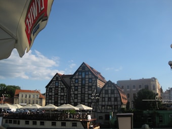 A quaint European street scene with distinctive timber-framed buildings and triangular roofs under a clear blue sky. Umbrellas with seating areas line the foreground, suggesting a caf&eacute; or outdoor dining setting. In the left corner, a large white and red umbrella with branding is visible, adding a touch of urban ambiance.