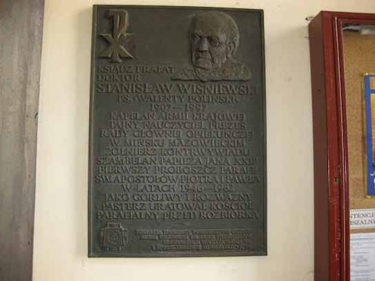 A bronze commemorative plaque mounted on a white wall. The plaque features raised text honoring a religious figure, with a portrait relief at the top right. Adjacent to the plaque is a bulletin board with a visible red frame.