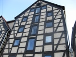 A traditional half-timbered building with dark wooden beams and white plaster walls. The structure is several stories tall, with multiple rectangular windows symmetrically placed on the facade. The steep pitched roof and the architectural style suggest a historic or European influence.