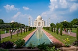 A majestic white mausoleum with a large dome and minarets stands in the center of a landscaped garden. The foreground features a long, reflective pool that leads the eye towards the structure, flanked by symmetrical rows of lush green trees and pathways where visitors are walking and gathering.