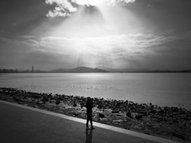 A tranquil seascape featuring a lone person walking along a paved pathway beside a rocky shore. The sky is partly cloudy, and beams of sunlight pierce through the clouds, casting a serene glow over the water. Distant mountains and a city skyline are visible in the background.