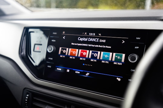 A car dashboard displays a digital infotainment system screen. The screen shows a radio interface with various channels like BBC R1, BBC R2, and BBC R6M. The current station playing is Capital DANCE with a song by Tall Paul. The interface includes options for radio, media, phone, and voice control, and the current time is displayed.