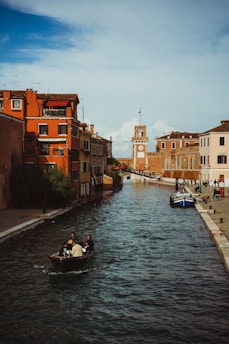 A picturesque canal winds through a historic European city, flanked by vibrant buildings with terracotta roofs. A small boat carries passengers along the waterway, while a few pedestrians walk along the cobblestone paths. In the distance, a tall clock tower rises against a partly cloudy sky, enhancing the old-world charm of the scene.