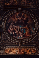 A richly detailed and dramatic classical artwork featuring mythological figures arranged in an ornate circular frame. The scene includes dynamic poses, flowing garments, and intricate detailing, all set against a vividly colored backdrop. The craftsmanship of the gold embellishments highlights the grandeur of the composition.