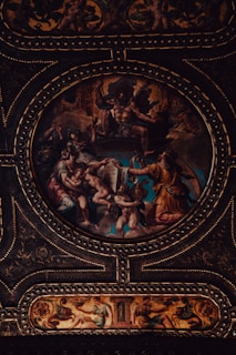 A richly detailed and dramatic classical artwork featuring mythological figures arranged in an ornate circular frame. The scene includes dynamic poses, flowing garments, and intricate detailing, all set against a vividly colored backdrop. The craftsmanship of the gold embellishments highlights the grandeur of the composition.