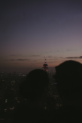 A silhouetted couple stands in the foreground, overlooking a cityscape at dusk. The skyline features numerous buildings, with a prominent lit structure in the center, possibly a famous skyscraper. The sky is transitioning from dusk to night, with a gradient of colors from dark blue to a lighter hue near the horizon.