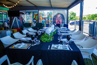 A long dining table is elegantly set for a meal, covered with a dark tablecloth and adorned with white flowers and greenery running down the center. Each place is prepared with plates, napkins, and black wine glasses. Surrounding the table are modern white chairs. In the background, colorful murals with abstract and pop-art designs are visible, featuring stylized faces in vibrant colors. The space is partially outdoors, covered by a roof with strings of lights visible above. Large windows provide a view of the outside, where greenery and a parked car can be seen.