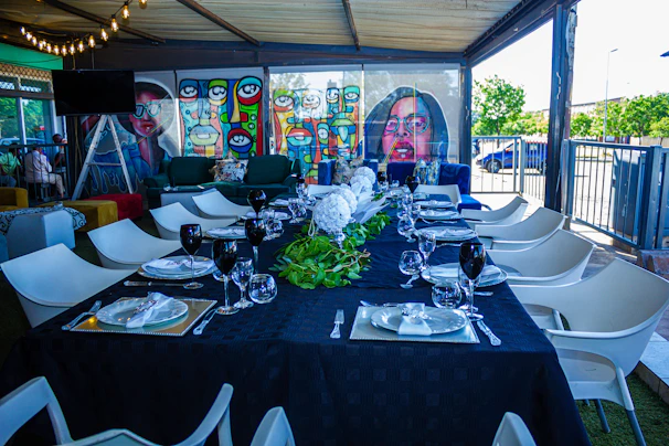 A long dining table is elegantly set for a meal, covered with a dark tablecloth and adorned with white flowers and greenery running down the center. Each place is prepared with plates, napkins, and black wine glasses. Surrounding the table are modern white chairs. In the background, colorful murals with abstract and pop-art designs are visible, featuring stylized faces in vibrant colors. The space is partially outdoors, covered by a roof with strings of lights visible above. Large windows provide a view of the outside, where greenery and a parked car can be seen.