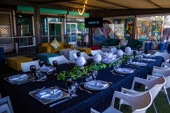 A beautifully set dining table is adorned with a dark tablecloth, elegant glassware, white plates, and white floral centerpieces. Greenery runs along the center of the table, adding a natural element. Surrounding the table are white chairs. In the background, there are colorful murals depicting abstract faces and geometrical patterns. Comfortable seating with vibrant cushions is placed against the wall, creating a cozy atmosphere. A screen mounted on the wall displays the word 'LALIGA'.