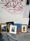 Several framed artworks and photographs are leaning against an exterior wall with red graffiti on it. The images include a black-and-white photograph of a person with a pram, a monochrome portrait, a colorful painting of the Eiffel Tower, and floral paintings. The wall is light-colored and slightly dirty, and the ground appears to be concrete with cracks and stains.