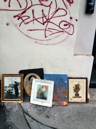 Several framed artworks and photographs are leaning against an exterior wall with red graffiti on it. The images include a black-and-white photograph of a person with a pram, a monochrome portrait, a colorful painting of the Eiffel Tower, and floral paintings. The wall is light-colored and slightly dirty, and the ground appears to be concrete with cracks and stains.