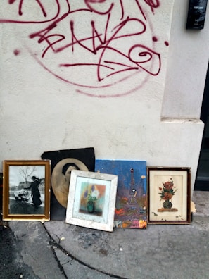 Several framed artworks and photographs are leaning against an exterior wall with red graffiti on it. The images include a black-and-white photograph of a person with a pram, a monochrome portrait, a colorful painting of the Eiffel Tower, and floral paintings. The wall is light-colored and slightly dirty, and the ground appears to be concrete with cracks and stains.