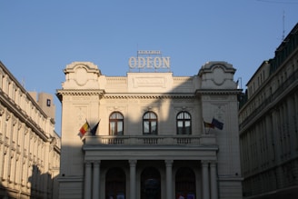 A neoclassical theatre building with ornate architectural details and columns at the entrance. The facade features symmetrical windows and flags are visible on either side. The lighting creates shadows on the structure, enhancing its detailed features.