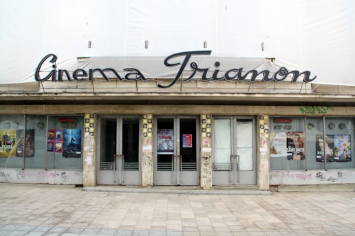 A vintage cinema facade featuring 'Cinema Trianon' in stylized lettering above the entrance. The building has closed doors and is adorned with various movie posters on either side. The exterior is slightly weathered, with graffiti seen on the lower areas and walls surrounding the entrance.