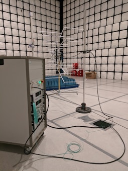 A room with a grid-patterned wall that resembles an anechoic chamber, containing various electronic equipment. There is a large antenna-like structure in the middle and several cables running across the floor connected to a control panel or server rack. Blue absorptive materials are placed around the antenna setup for sound or signal management. A stack of red crates and a cardboard box are in the background.
