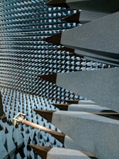 A space filled with large, dark gray, wedge-shaped foam structures that line the walls, ceiling, and floor, creating a visually striking pattern. This is likely an anechoic chamber designed to absorb sound or electromagnetic waves. A metal arm or fixture protrudes from one side, possibly holding equipment.