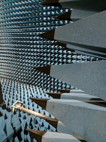 A space filled with large, dark gray, wedge-shaped foam structures that line the walls, ceiling, and floor, creating a visually striking pattern. This is likely an anechoic chamber designed to absorb sound or electromagnetic waves. A metal arm or fixture protrudes from one side, possibly holding equipment.