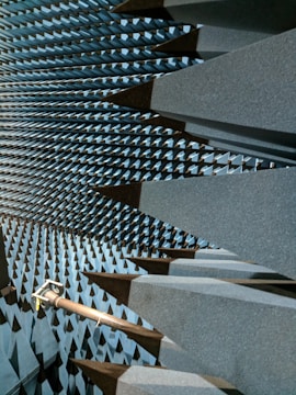 A space filled with large, dark gray, wedge-shaped foam structures that line the walls, ceiling, and floor, creating a visually striking pattern. This is likely an anechoic chamber designed to absorb sound or electromagnetic waves. A metal arm or fixture protrudes from one side, possibly holding equipment.