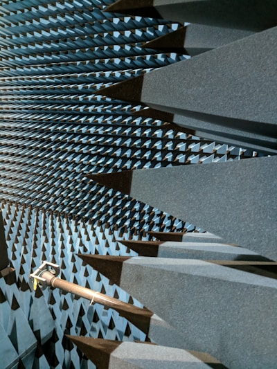 A space filled with large, dark gray, wedge-shaped foam structures that line the walls, ceiling, and floor, creating a visually striking pattern. This is likely an anechoic chamber designed to absorb sound or electromagnetic waves. A metal arm or fixture protrudes from one side, possibly holding equipment.