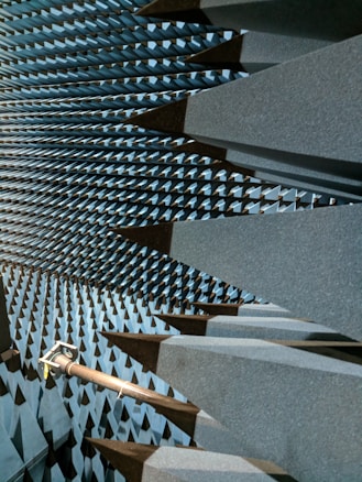 A space filled with large, dark gray, wedge-shaped foam structures that line the walls, ceiling, and floor, creating a visually striking pattern. This is likely an anechoic chamber designed to absorb sound or electromagnetic waves. A metal arm or fixture protrudes from one side, possibly holding equipment.
