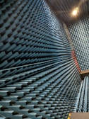 An interior space with walls covered in a dense arrangement of blue, pyramid-shaped acoustic foam panels arranged in a uniform pattern. The perspective creates a visual effect of depth and texture, with lighting that casts shadows across the surfaces.