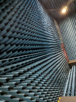 An interior space with walls covered in a dense arrangement of blue, pyramid-shaped acoustic foam panels arranged in a uniform pattern. The perspective creates a visual effect of depth and texture, with lighting that casts shadows across the surfaces.
