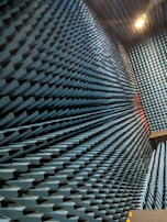 An interior space with walls covered in a dense arrangement of blue, pyramid-shaped acoustic foam panels arranged in a uniform pattern. The perspective creates a visual effect of depth and texture, with lighting that casts shadows across the surfaces.