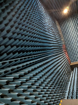 An interior space with walls covered in a dense arrangement of blue, pyramid-shaped acoustic foam panels arranged in a uniform pattern. The perspective creates a visual effect of depth and texture, with lighting that casts shadows across the surfaces.