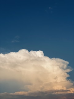 A large, fluffy cloud with a bright white top is contrasted against a deep blue sky. The cloud's edges are highlighted by sunlight, creating a dynamic interplay of light and shadow.