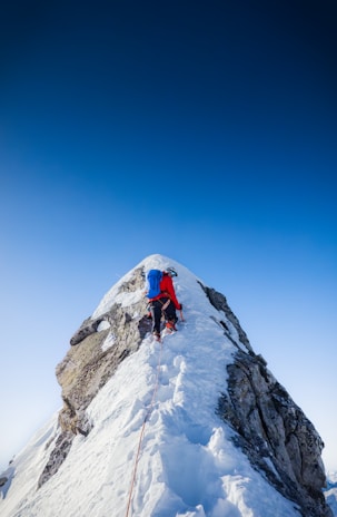 A climber wearing a red jacket and a blue backpack uses ice axes and crampons to ascend a steep, snowy mountain ridge. The sky is clear and blue, creating a striking contrast against the white snow.