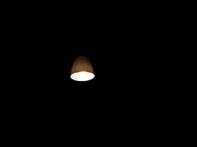 A well-lit lamp emits a soft glow, contrasting sharply with the surrounding darkness. The light source is encased in a simple, brown lampshade that diffuses the light gently.