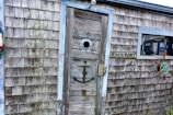 A weathered wooden shack with a rustic appearance, featuring a weathered door with a circular window. An anchor is mounted on the door. There is a sign reading 'ROCKPORT' next to a window with a fishing net and a wooden steering wheel decoration.